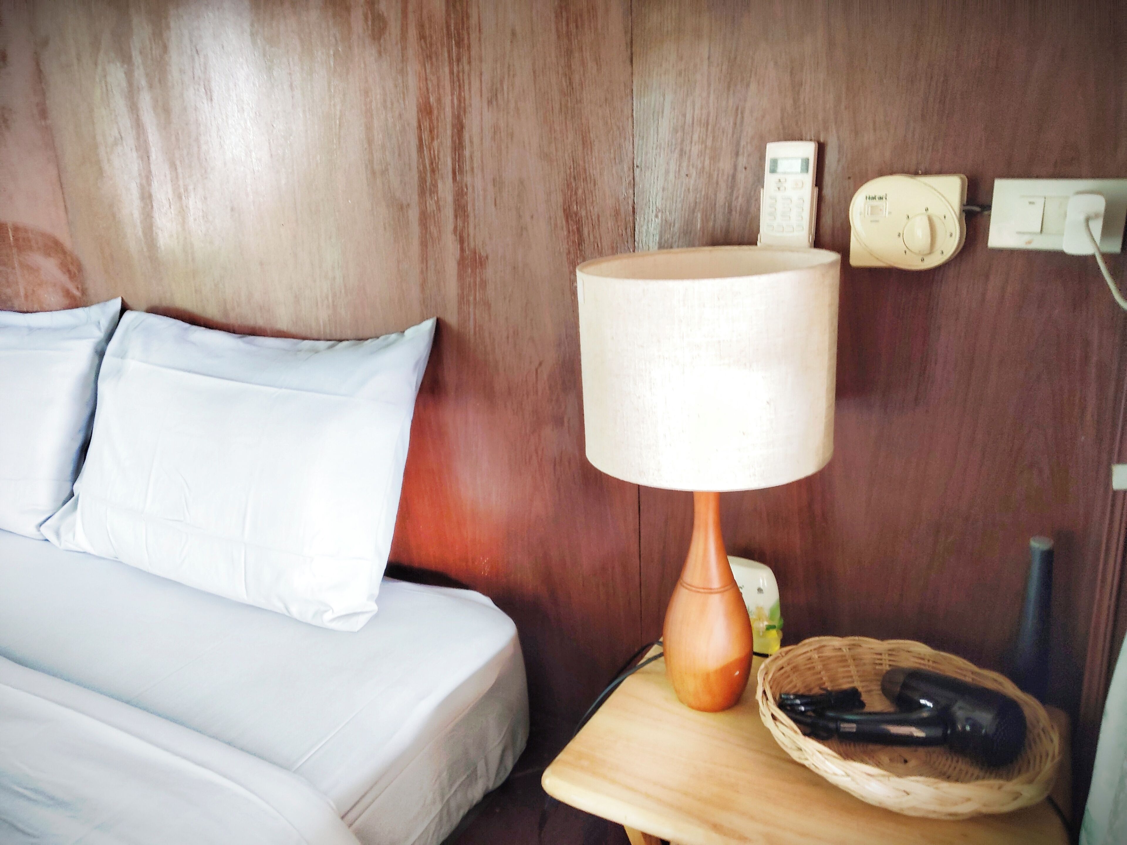 Un lit en bois avec une tête de lit, recouvert d’une literie blanche, agrémenté d’une lampe de chevet, d’un téléphone et d’un panier contenant un objet noir, le tout posé sur une table de chevet en bois.