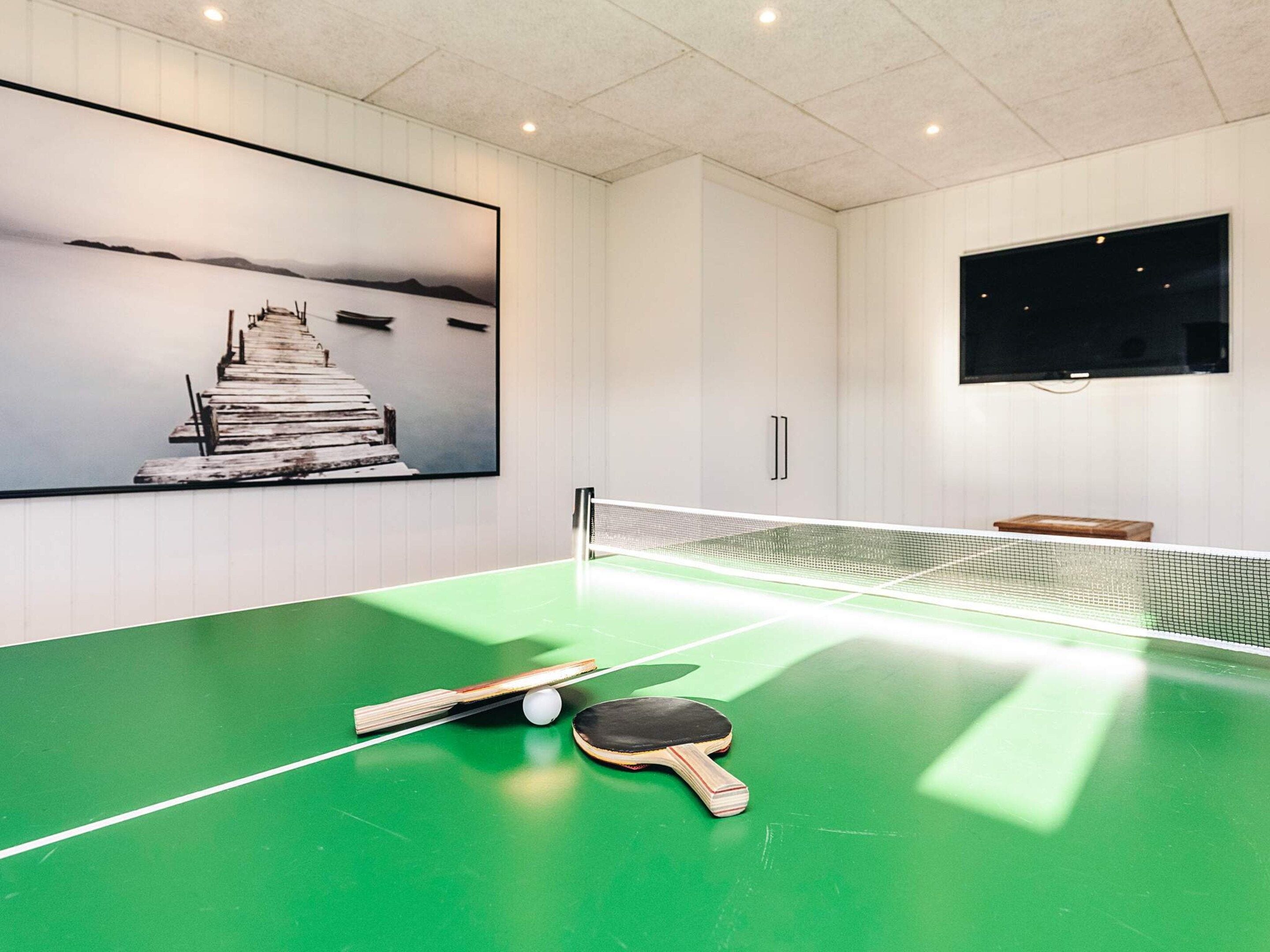 A ping pong table with paddles and a ball, a painting of a pier, and a TV on the wall.