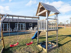 A playground with swings and a slide, a house with a dark exterior, and a clear sky. - 12 Person Holiday Home in Albaek (Aalbaek)