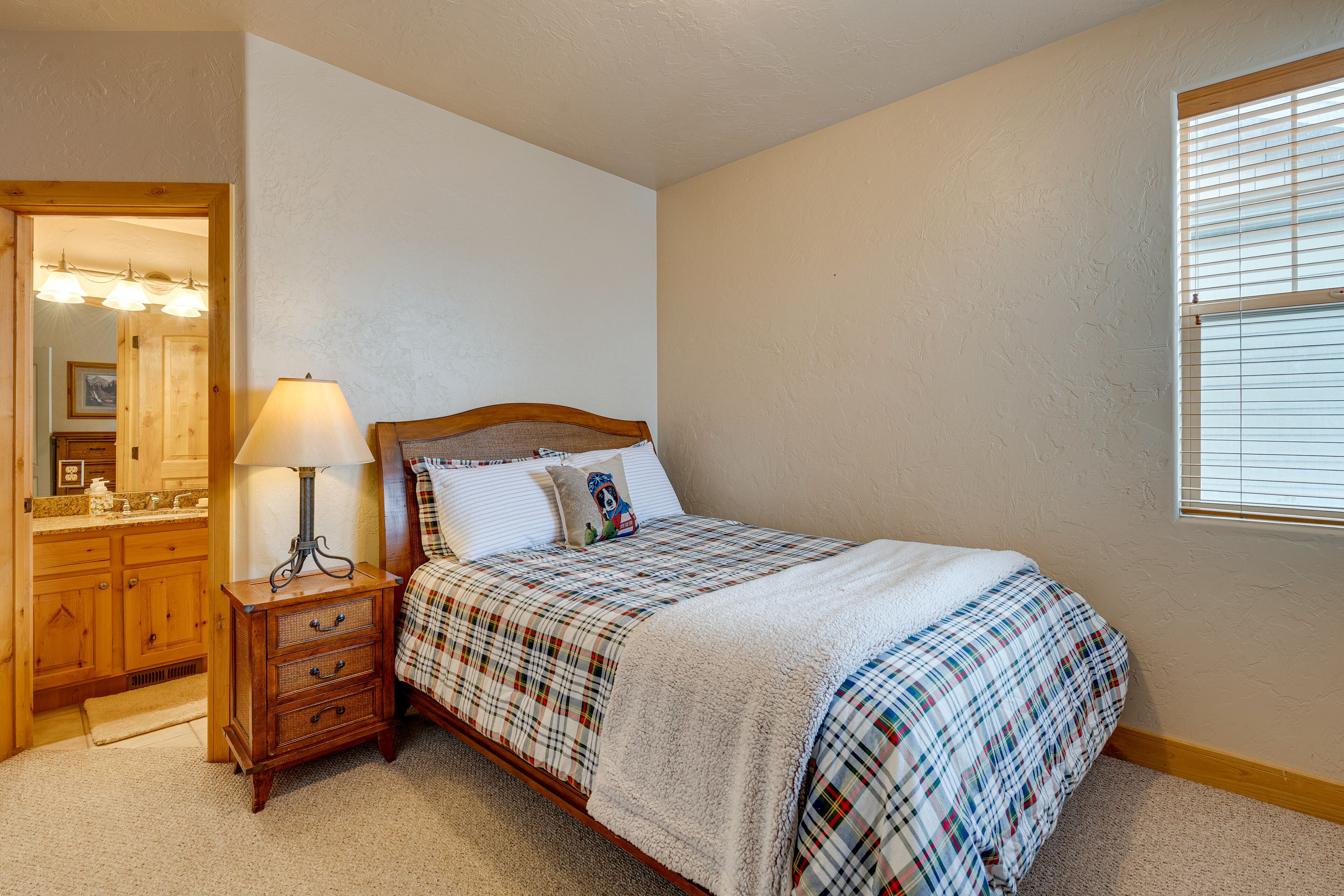 A bedroom with a bed, a nightstand, a lamp, a dresser, and a window with blinds.