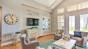 A living room with a clock, a flat-screen TV, a decorative wall with a quote, a wooden coffee table, and two armchairs. - Waterfront Lake Tawakoni Home w/ Fire Pit! (Quinlan)