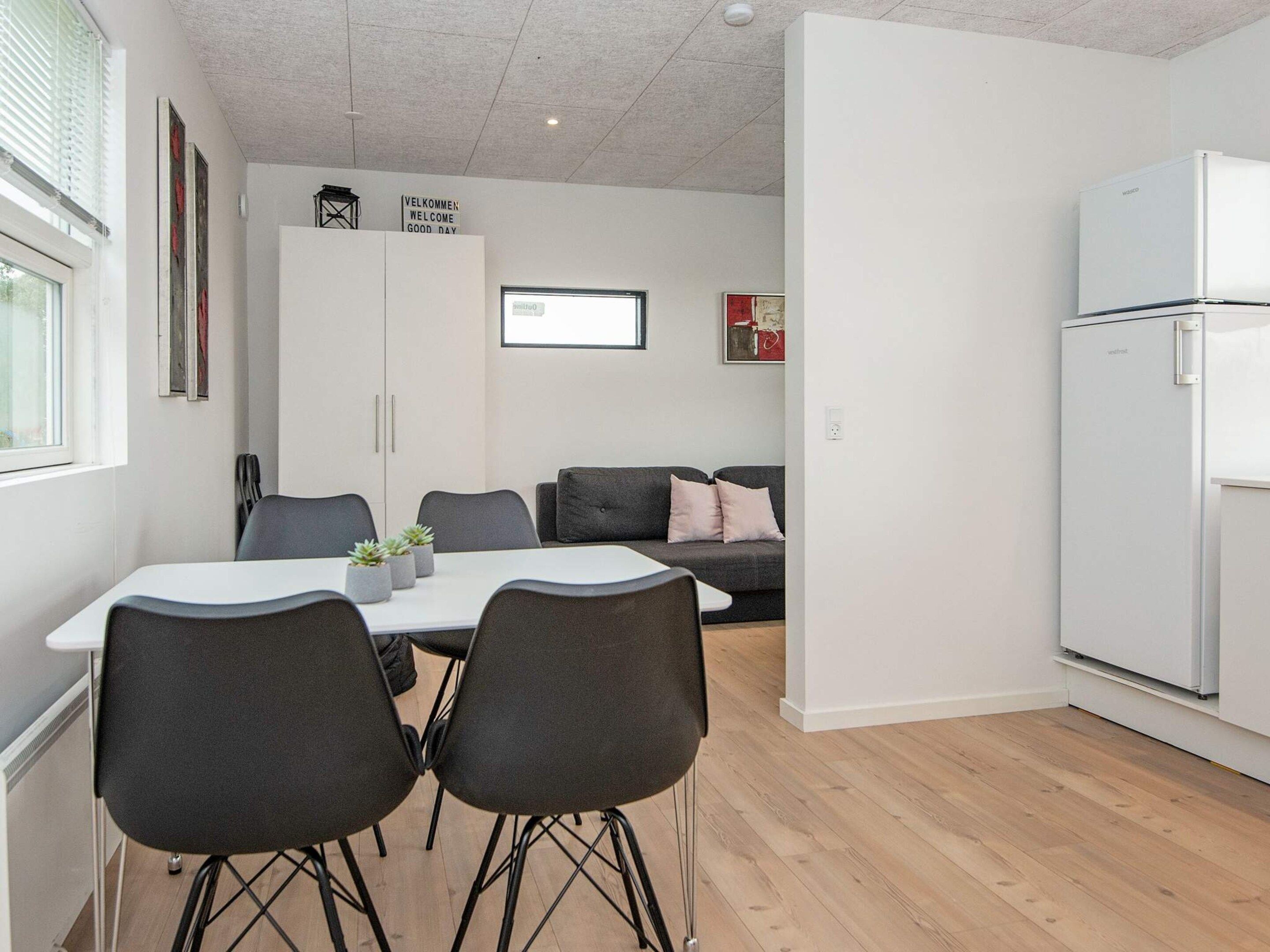 A modern kitchen with a white refrigerator, a dining table with four chairs, and a grey sofa in the background.