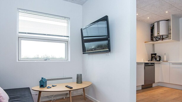 A compact room with a bed, a small table, a TV, a radiator, and a kitchen area with a dishwasher. - 5 Person Holiday Home in Ansager (Ansager)