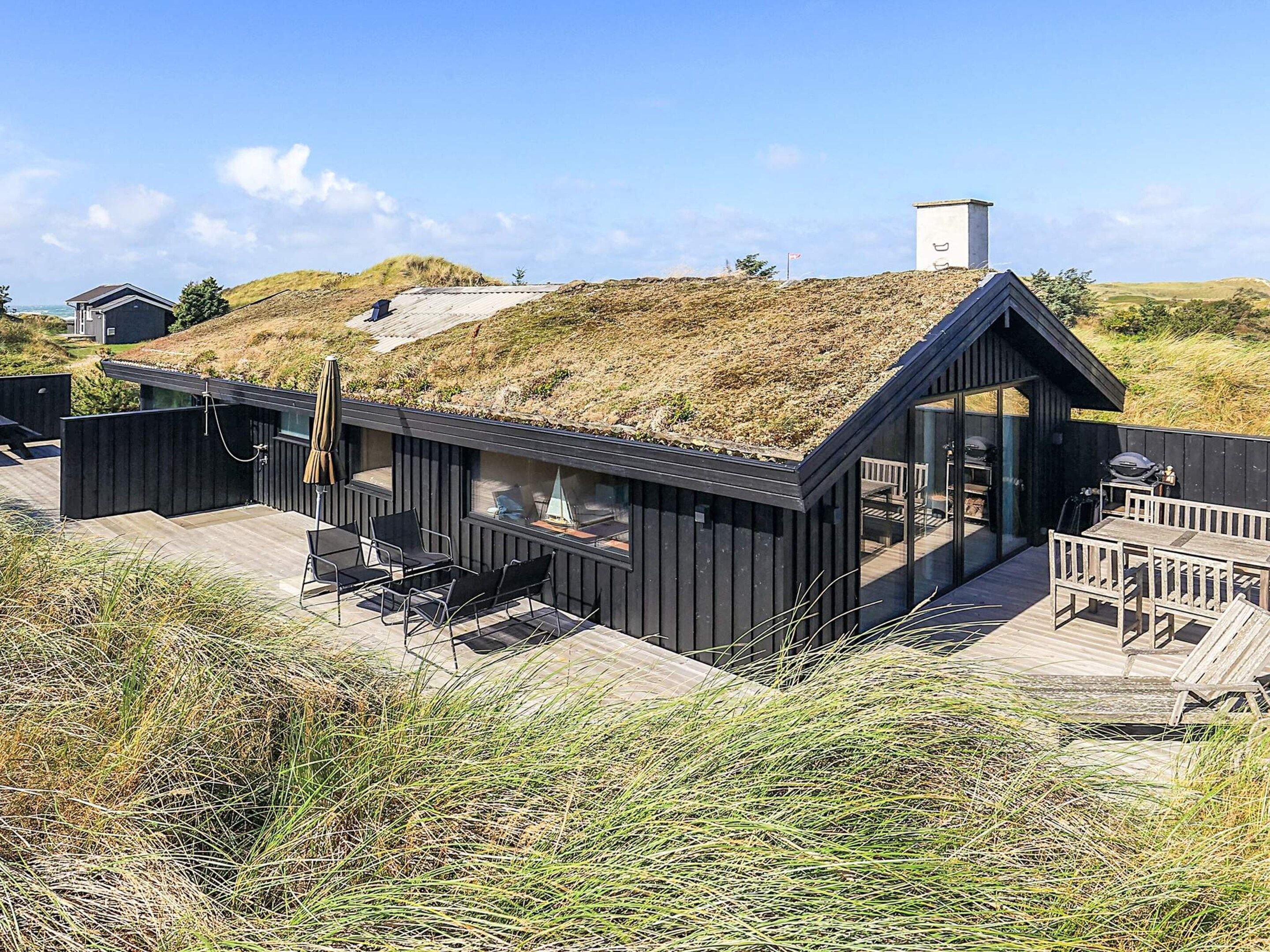 Una casa moderna frente a la playa, con techo plano, mobiliario exterior y vistas a las dunas.