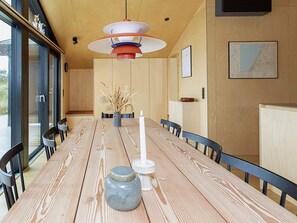 Uma sala de jantar com uma mesa de madeira comprida, cadeiras pretas, um vaso com plantas secas, uma vela e um pendente.