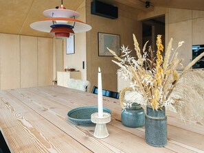 Una mesa de comedor de madera con una vela blanca, dos jarrones de cerámica con plantas secas y una lámpara colgante moderna.