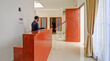 A hotel reception area with a wooden reception desk, a staircase, and a view of the outside through a window with curtains.
