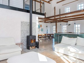 Ampla sala de estar com lareira, sofás brancos, mesa de jantar e cozinha com bancada verde.
