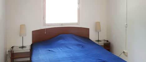 A single bed with a blue bedspread in a room with white walls and a window.