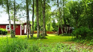 Une cabane en bois rouge avec une porte blanche, entourée d’un jardin luxuriant et verdoyant et de grands arbres.