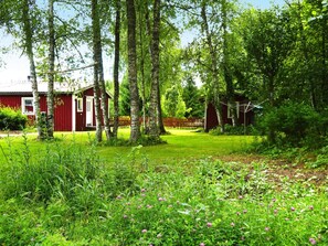 Une cabane en bois rouge avec une porte blanche, entourée d’un jardin luxuriant et verdoyant et de grands arbres.