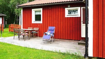 A red wooden building with a tiled roof, a small window, and a door. There is a wooden table and chairs outside, and a blue lounge chair on the porch.