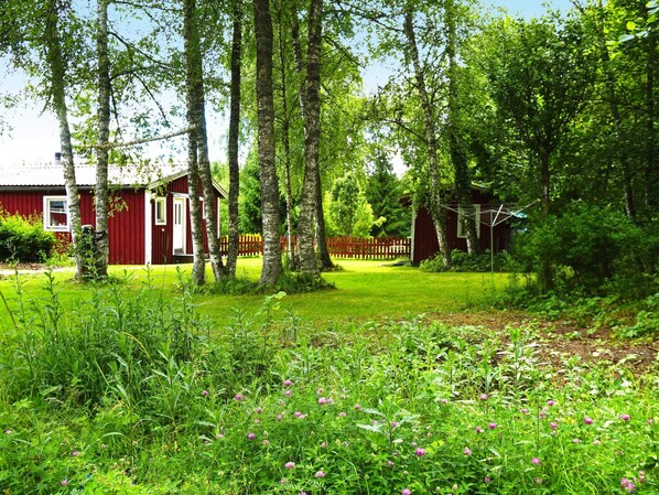 A red wooden cabin with a white door, surrounded by a lush green garden and tall trees. - 9 Person Holiday Home in Hallstavik (Hallstavik)