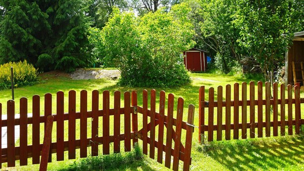 Une clôture en bois dans un jardin, avec un petit abri de jardin et un feuillage dense en arrière-plan.
