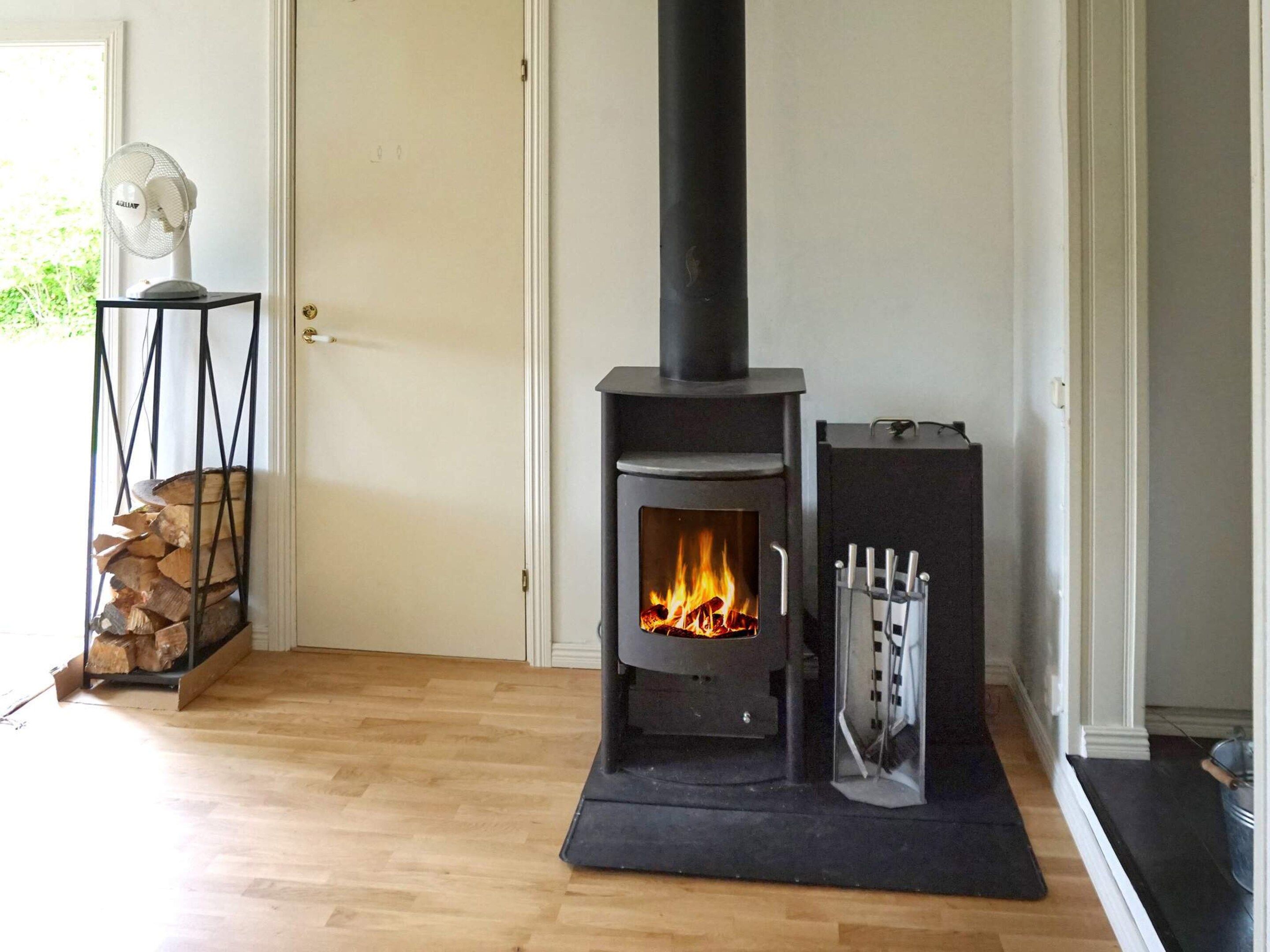 A room with a wood-burning stove, a door, a fan, and a stack of firewood.