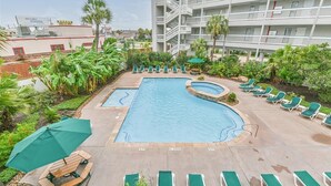 A hotel pool area with multiple lounge chairs and umbrellas, surrounded by greenery and a multi-story building.