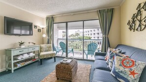 A living room with a sofa, chairs, a TV, a balcony with chairs, and a view of buildings.