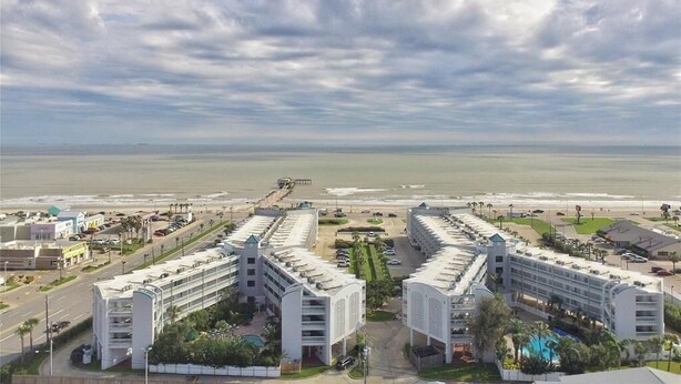 A coastal hotel with a symmetrical layout, featuring multiple floors and balconies, situated near a beach with a clear view of the ocean.