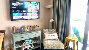 A room with a television displaying a sports event, a chair with a cushion, a small table, a basket, and a window with curtains.