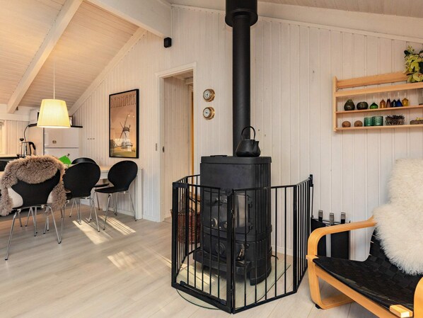 A cozy room with a black wood-burning stove, a dining area with chairs, a wooden shelf with decorative items, and a framed picture on the wall.
