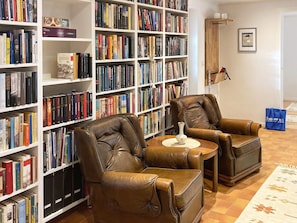 Um quarto com duas poltronas de couro, uma mesinha redonda e uma estante repleta de livros.
