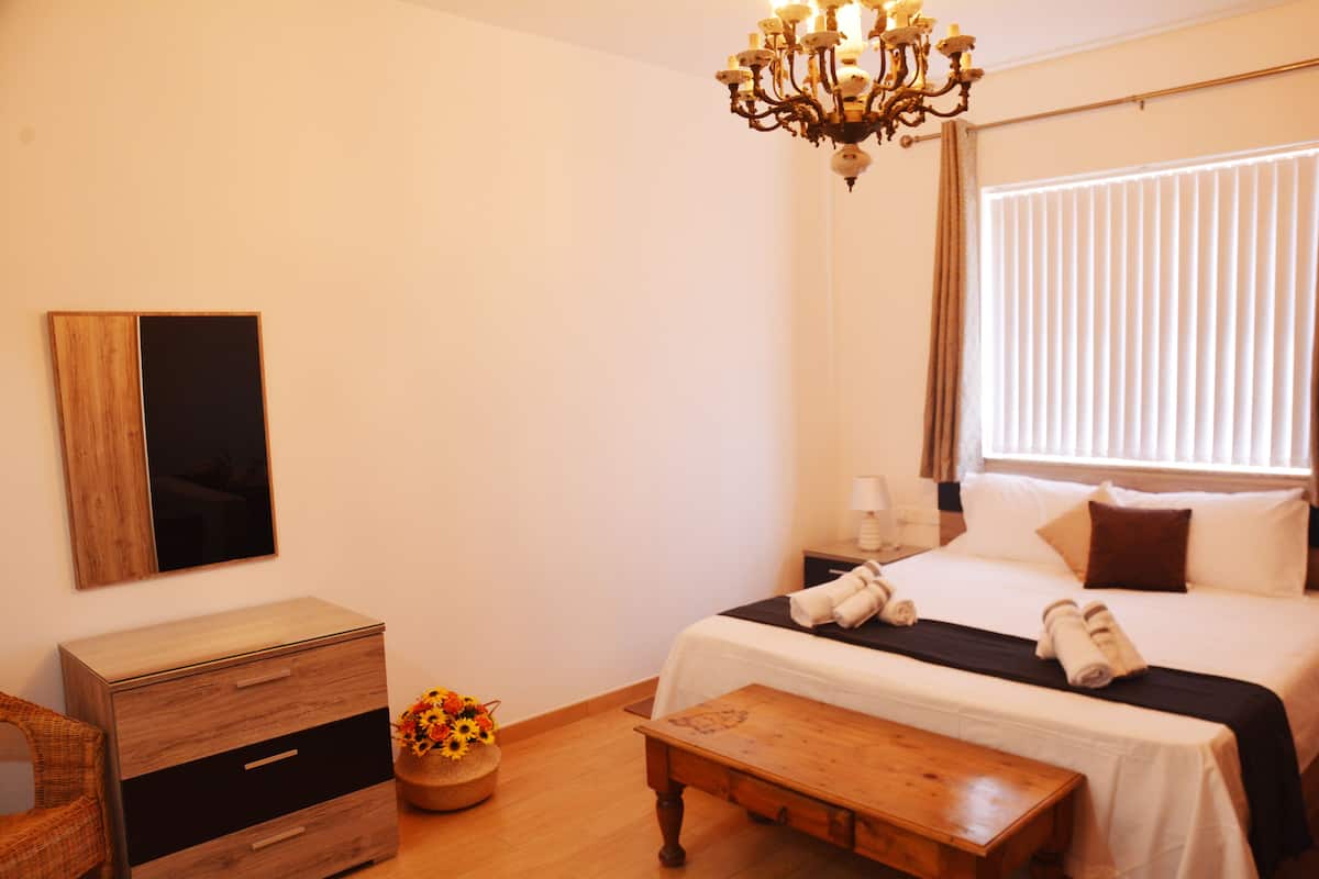 A hotel room with a wooden dresser, a bedside table, a chandelier, a bed with pillows, a nightstand, and a window with blinds.