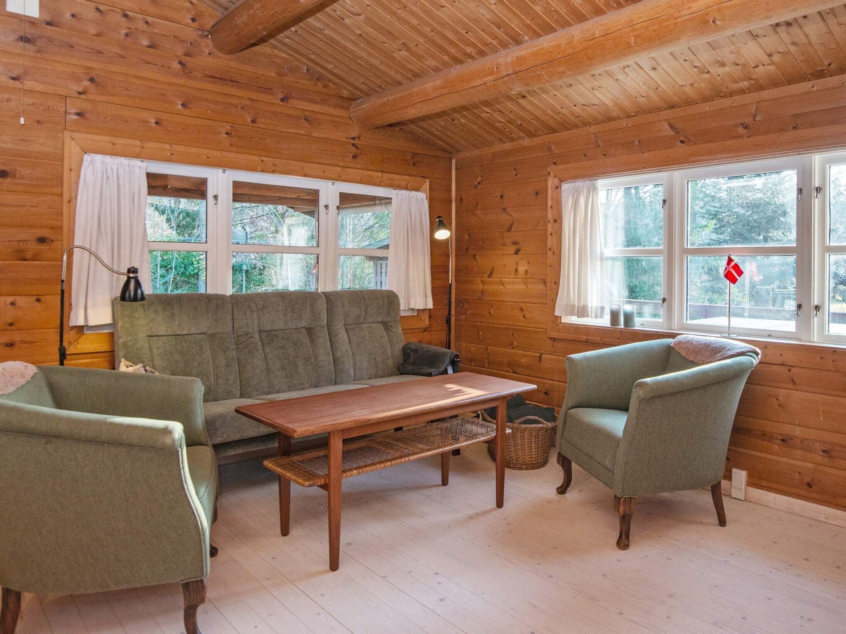 Ein gemütliches Blockhaus-Interieur mit einer Couch, zwei Sesseln, einem Holztisch und großen Fenstern mit Vorhängen.