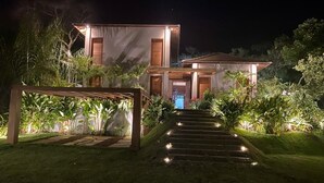 A modern house with a well-lit entrance, a staircase leading to the main door, and a garden area with various plants. - House with beautiful pool and private cook! (Mata de São João)