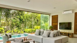 A modern living room with a large grey sectional sofa, a round coffee table, and a TV on the wall. There is a large window with a view of a swimming pool and lush greenery outside. - House with beautiful pool and private cook! (Mata de São João)