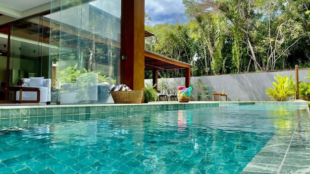 A swimming pool with clear water, surrounded by a modern house with large glass windows, a covered outdoor seating area, and lush greenery. - House with beautiful pool and private cook! (Mata de São João)