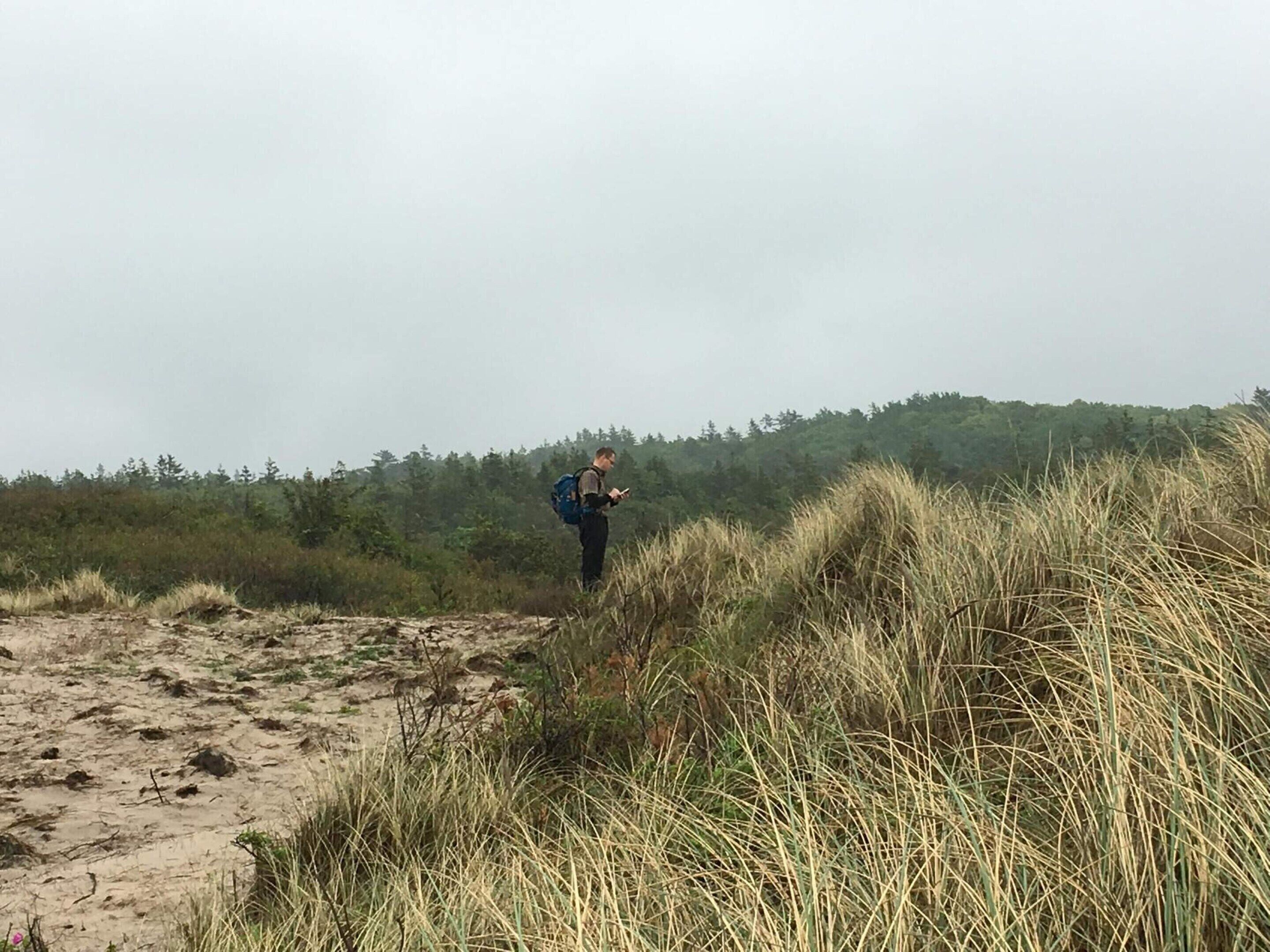 Iemand staat in een natuurlandschap met zand en gras, en draagt een rugzak.