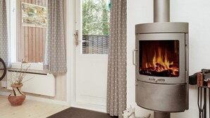 A room with a wood-burning stove, a door with curtains, a window with a view of greenery, and a potted plant. - 5 Person Holiday Home in Frederiksvaerk (Frederiksvaerk)