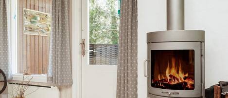 A room with a wood-burning stove, a door with curtains, a window with a view of greenery, and a potted plant.
