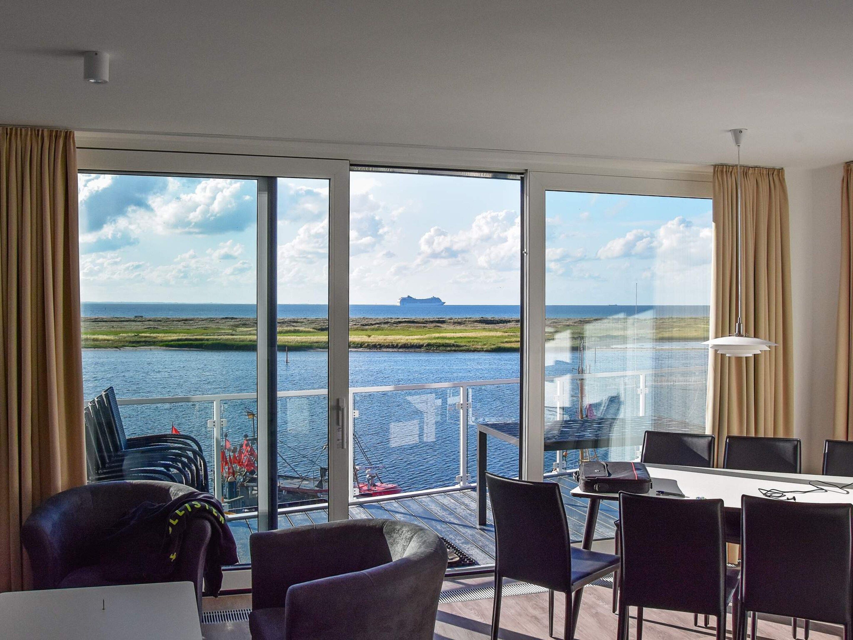 A modern room with a large window overlooking a body of water and a distant island. There is a table with chairs and a laptop.