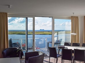 A room with a large window overlooking a body of water and a distant island. There is a table with chairs and a laptop on it.