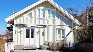 Ett tvåvåningshus med vit träfasad, en veranda framför, en liten trappa och en inhägnad trädgård.