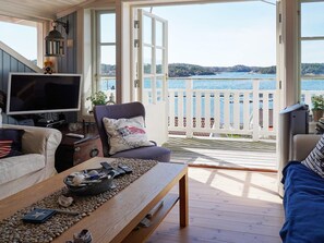 A cozy living room with a television, a coffee table with decorative items, a comfortable chair, and a view of a lake through open doors. - 4 Star Holiday Home in Tanumshede (Tanum)