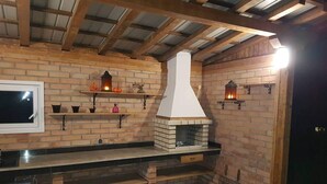 A rustic kitchen with a brick wall, wooden beams, a built-in brick oven, and a shelf with decorative items.