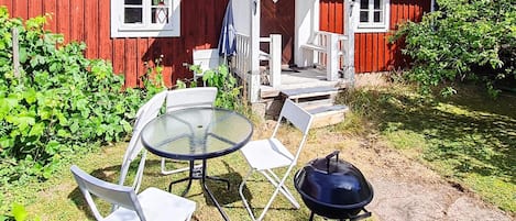 A red wooden house with a porch, a glass table with chairs, and a barbecue grill.