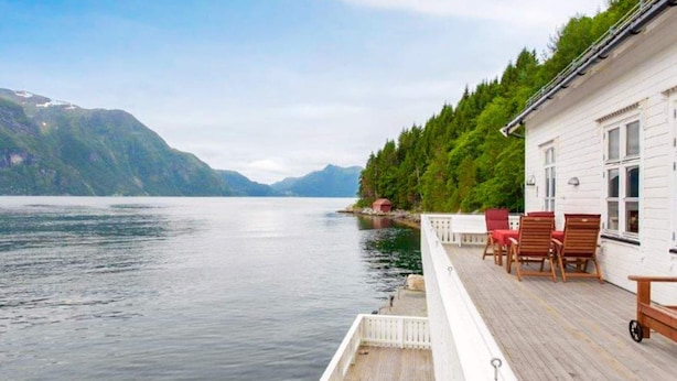 A wooden deck with chairs, a white building with windows, and a lake with mountains in the background. - 8 Person Holiday Home in Orskog (Ålesund)
