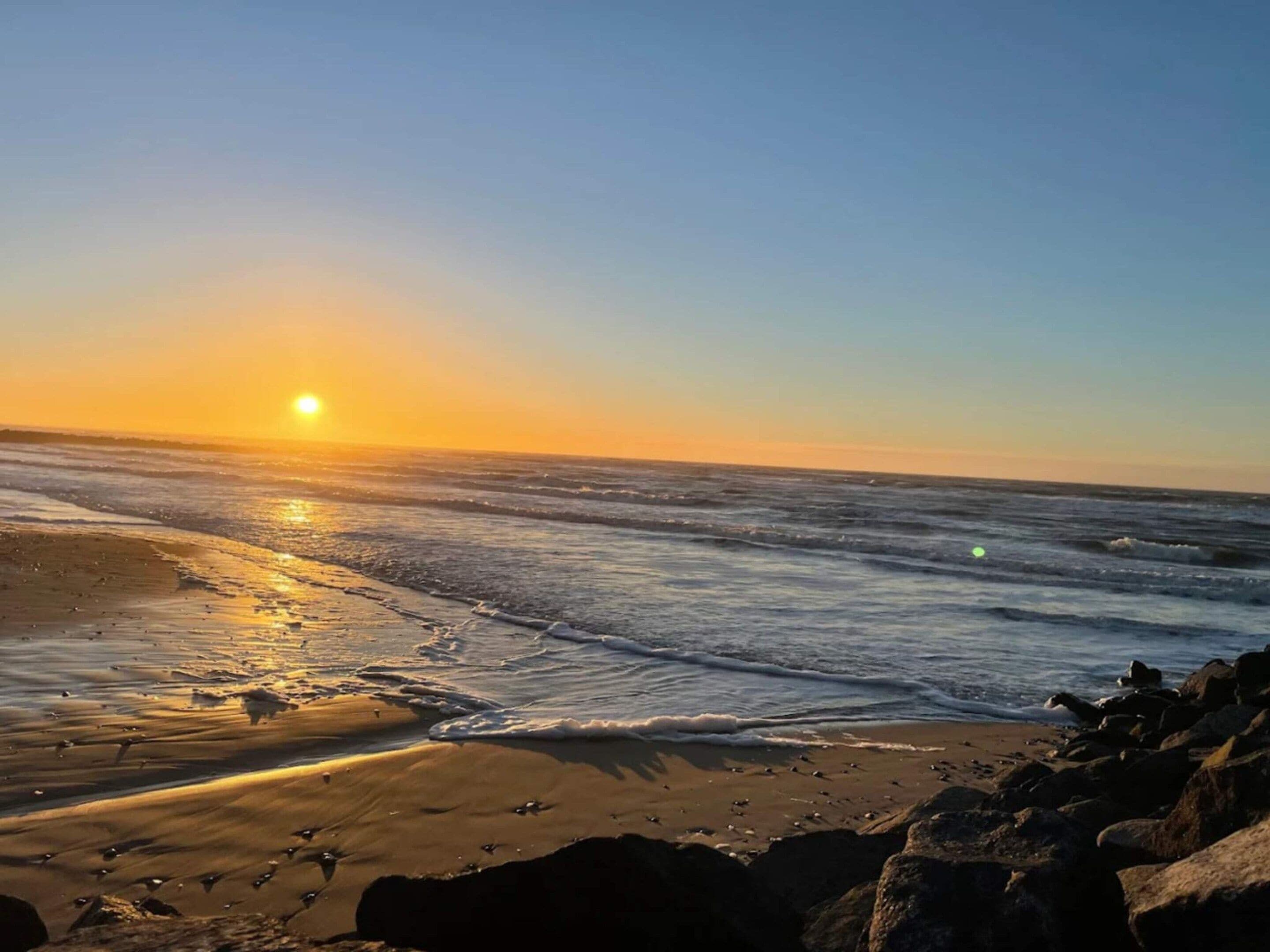 Et strandbillede med solnedgang over havet, bølger der forsigtigt slår ind mod kysten, og sten i forgrunden.