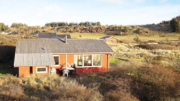 Una cabaña de madera con techo inclinado, un patio con muebles de exterior y vistas a un campo cubierto de césped y árboles al fondo.