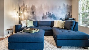 A living room with a sectional sofa, ottoman, and a forest-themed wall mural.