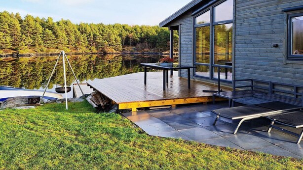 A modern cabin with a deck overlooking a lake, surrounded by dense forest.