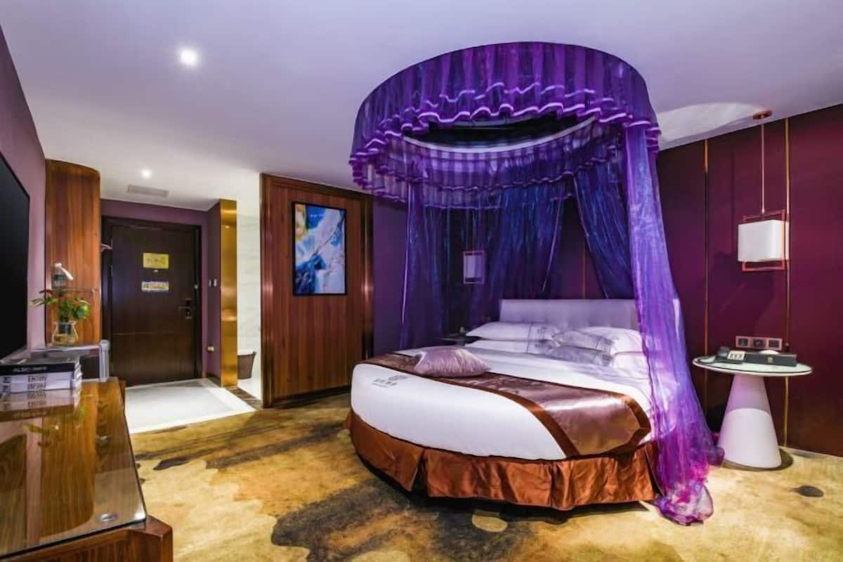 A hotel room with a bed, a desk, a chair, a small table, and a wall-mounted TV.