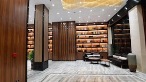 A modern lobby with a marble floor, a seating area with sofas and tables, and a wall of illuminated shelves.