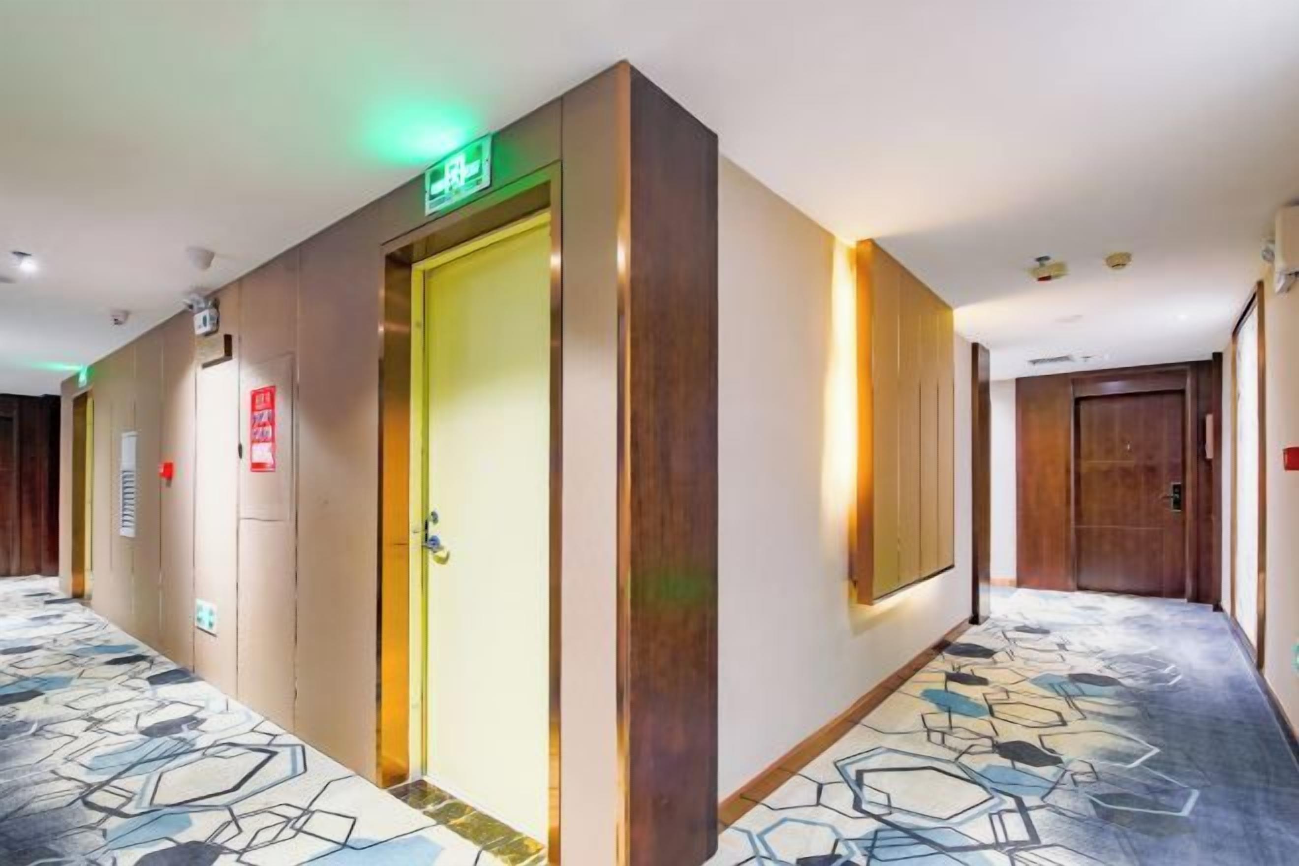 A hotel hallway with a patterned carpet, a door with an emergency exit sign, and a wall-mounted light fixture.