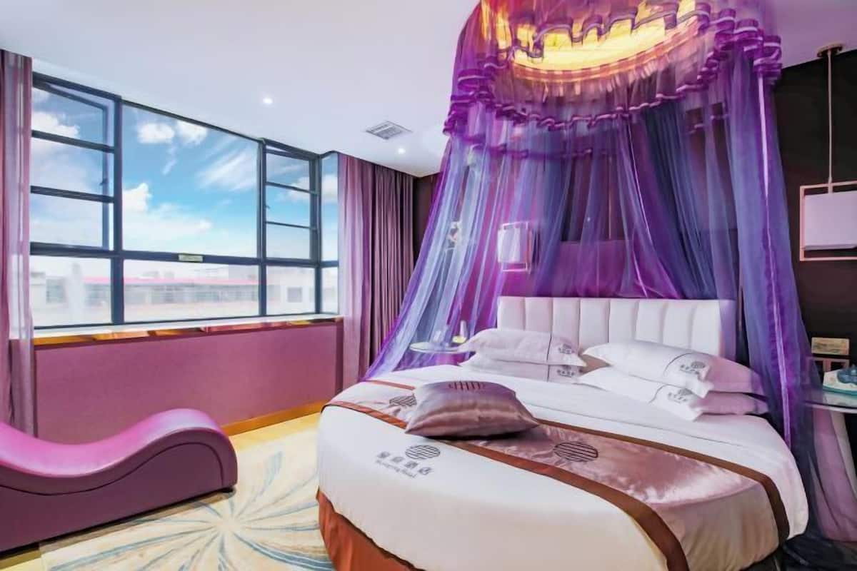 A hotel room with a large window, a bed with a canopy, and a pink chair.