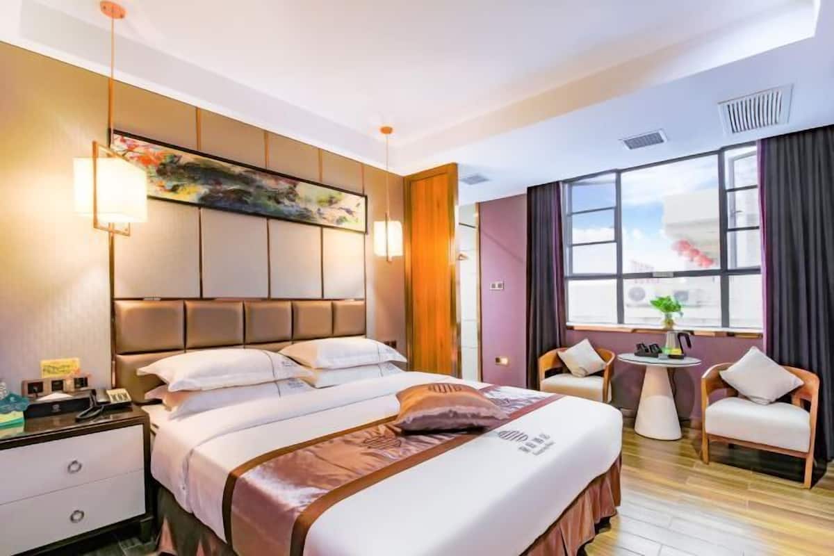 A hotel room with a large bed, a bedside table, a small round table, two chairs, a window with curtains, and a painting on the wall.