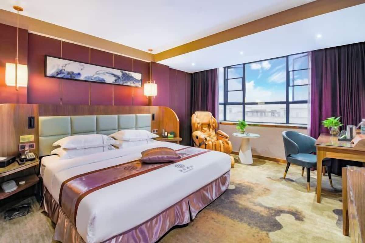 A hotel room with a large bed, a massage chair, a desk, and a view of the city.